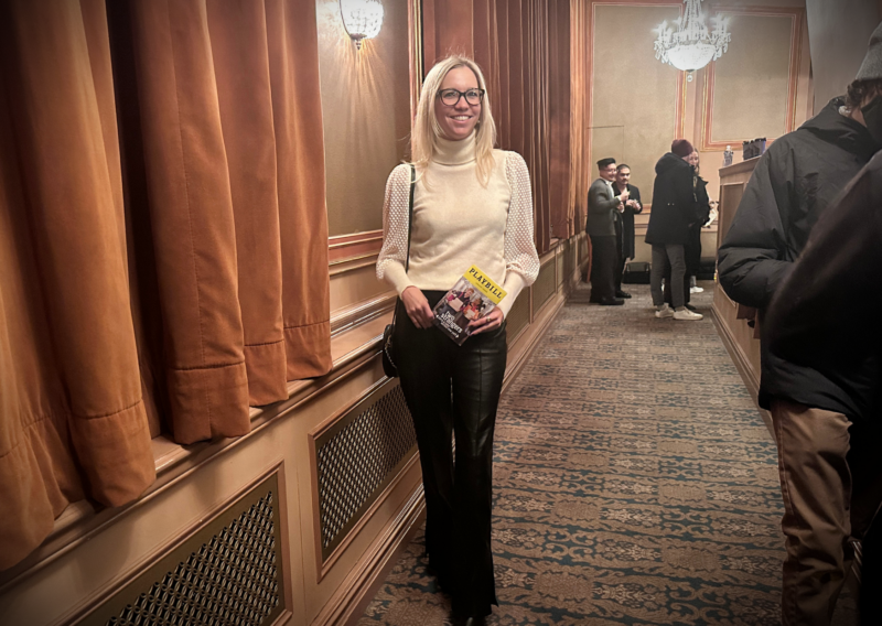 Ally at the theater seeing broadway show two strangers (carrying a cake across new york) -wearing leather trousers and a cream cashmere sweater holding a playbill at the Longacre Theatre